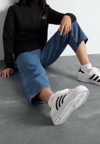 Svart zip-up sweatshirt med liten logotyp, blå vida jeans med råa kanter, och vita sneakers med svarta ränder, sittande på en grå yta.