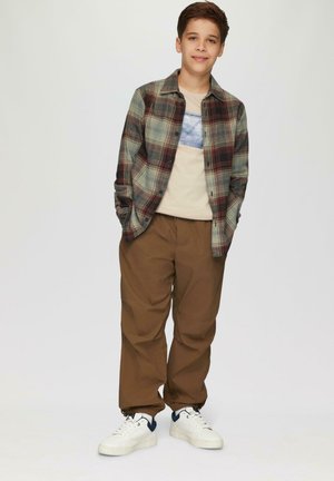 Een geruite overhemd met groene, rode en bruine tinten, een beige graphic t-shirt, bruine baggy broek en witte sneakers met marineblauwe accenten.