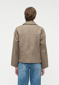 Taupe faux suedejakke med en avslappet passform, store skuldersømmer og detalj på rygggjelden, kombinert med lyseblå denimjeans.