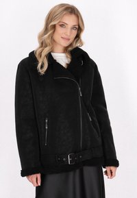 Blonde Frau trägt eine schwarze Schaffelljacke mit Rei�ßverschlusstaschen, einem Schnallen-Gürtel und einem weißen Oberteil darunter, steht vor einem schlichten Hintergrund.