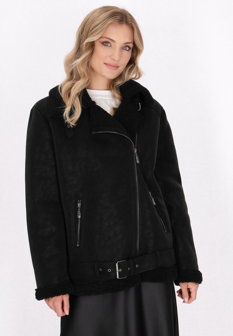 Blonde Frau trägt eine schwarze Schaffelljacke mit Reißverschlusstaschen, einem Schnallen-Gürtel und einem weißen Oberteil darunter, steht vor einem schlichten Hintergrund.