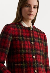 Sarkanas un zaļas paklājveida žaketes, kas izgatavota no teksturētas vilnas, ar noapaļotu apkakli, zeltītiem ziedu pogām un piegulošu siluetu.