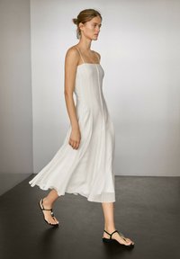 Weißes ärmelloses Kleid mit quadratischem Ausschnitt, fließender A-Linie Silhouette und vertikalen Nähten. Kombiniert mit schwarzen Riemchensandalen.
