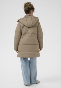 Beige pufferjakke med hætte, der har syede paneler og et bælte i taljen, båret over lyseblå vide jeans. Glat tekstur og afslappet design.
