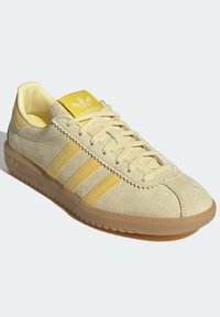 Beige mokkasneakers med gule detaljer, med tre sidestreker, snøring og en teksturert gummisåle. Rund tå-design.