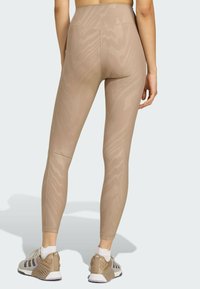 Beige Leggings mit hoher Taille und subtiler gemusterter Struktur, körpernah und knöchellang, kombiniert mit beigen und grauen Sportschuhen.