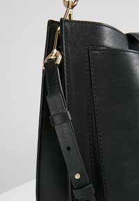 Czarna skórzana torba o konstrukcji z wyraźną strukturą, z detalami szycia i odpinanym paskiem przymocowanym złotym okuciem.