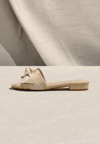 Μπεζ σανδάλι τύπου slip-on με υφαντή υφή στη σόλα και στο λουράκι, διακοσμημένο με μεγάλο στριμμένο φιόγκο στο μπροστινό επάνω μέρος.