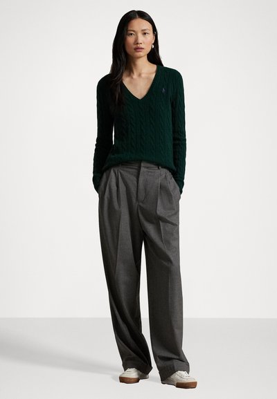 Groene kabelgebreide trui met een V-hals ontwerp, gecombineerd met grijze geplooide broek en witte sneakers. Het outfit heeft een ontspannen pasvorm en een zachte textuur.