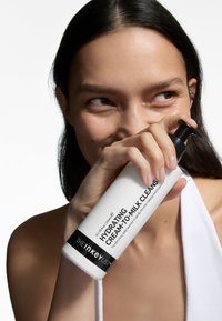 Hvid cylindrisk flaske med sort pumpehat, mærket "Hydrating Cream-to-Milk Cleanser." Glat tekstur, klart produkt synlighed, minimalistisk design.
