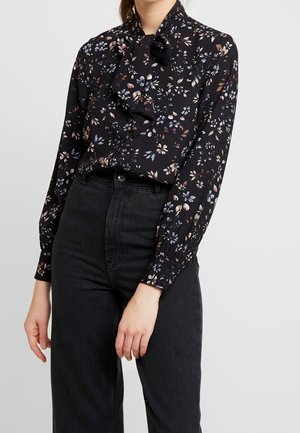 Nainen pukeutuneena mustaan kukkakuviolliseen pitkähihainen paitaan, jossa on solmittu rusetti kaulassa, ja korkeavyötäröisiin mustiin farkkuihin, seisoo yksiväristä taustaa vasten.