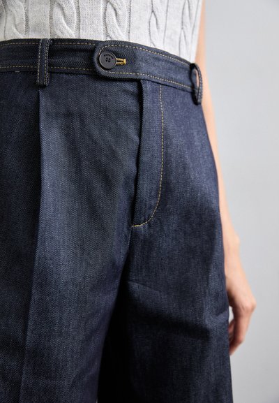Pantalons en denim foncé avec une taille haute, des plis et des coutures jaunes contrastantes. Un bouton rond sécurise la taille.