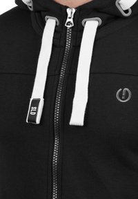 Schwarzer Hoodie mit weißen Kordelzügen, durchgehendem Reißverschluss und kleinem Logo auf der Brust. Hergestellt aus glattem Stoff mit weicher Textur.