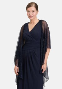 Donna che indossa un abito avvolgente blu navy con scollo a V, con un leggero e scintillante scialle blu navy che le ricade sulle spalle, in piedi davanti a uno sfondo bianco.