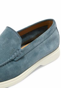 Mocassino in suede blu con una texture liscia, cuciture dettagliate e suola in gomma bianca. Nome del marchio impresso di lato.