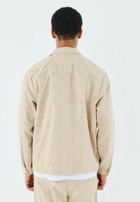 Chaqueta beige con un sutil patrón repetido de "PXP", cuello, mangas largas y puños abotonados, vista desde la parte posterior. Textura suave, corte relajado.