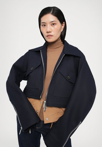 Chaqueta corta de color marino con mangas anchas, que cuenta con bolsillos frontales, cierre de cremallera y un forro interior en color marrón claro. Textura de tela suave.