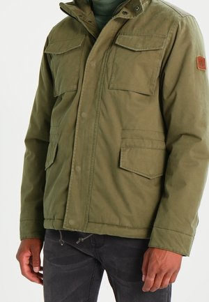 Homme portant une veste vert olive avec plusieurs poches à rabat, debout devant un fond clair uni.