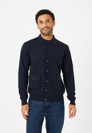 Man draagt een donkerblauwe, met knopen gesloten cardigan met voorzakken en een blauwe spijkerbroek, staand tegen een witte achtergrond.