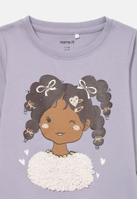 Lilla baby skjorte med en blinkende jenteillustrasjon, krøllete hår bundet med hvite sløyfer, hjerteformede hårklemmer, og en teksturert hvit topp.