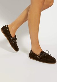 Brown suede moccasin-style shoes with gold stud accents, lace-up detail, and a flexible sole, featuring a soft inner lining.