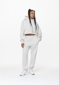 Jonge vrouw met lange vlechten, gekleed in een crèmekleurige cropped hoodie, bijpassende sweatpants en witte sneakers, staande tegen een witte achtergrond.