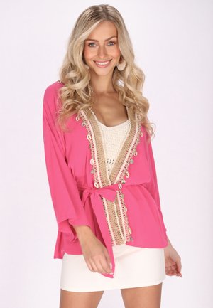 Cardigan stil kimono de culoare roz, cu mâneci largi, detaliu de croșetat crem și accente decorative. Legat la talie, asortat cu o fustă albă.