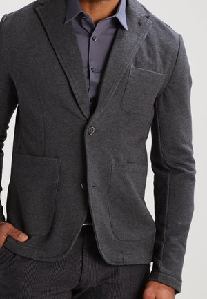 Man wearing dark grey blazer over buttoned dark grey shirt, paired with dark checkered pants, hand resting in pocket.