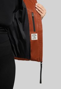 Giacca esterna marrone con fodera interna nera, dotata di una tasca con zip e un'etichetta in tessuto con testo. Texture liscia e design elegante.