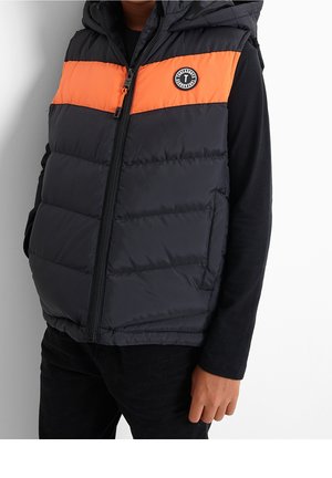 Enfant portant un gilet matelassé noir et orange avec un logo circulaire sur la poitrine par-dessus un t-shirt noir à manches longues.
