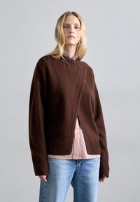 Rjav volnen pulover z ohlapnim krojem, ki ima ovit dizajn in rebraste manšete, oblečen čez rdečo in belo prugasto srajco s kragnico.