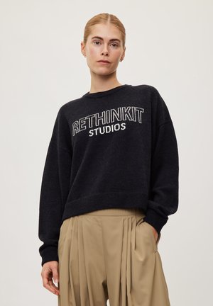Femme aux cheveux blonds porte un pull noir "RETHINKIT STUDIOS" et un pantalon beige plissé, debout les mains dans les poches devant un fond clair.