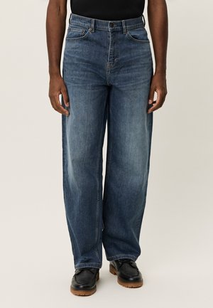 Άνδρας που φοράει μπλε τζιν με φαρδιά μπατζάκια και μαύρα δερμάτινα παπούτσια με κορδόνια, στέκεται μπροστά σε απλό ανοιχτόχρωμο φόντο.