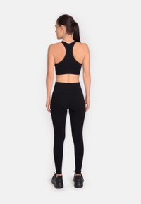 Sieviete stāv ar muguru pret priekšpusi, tērpta melnā sporta krūšturī ar racerback, augsto vidukli melnās legingos un melnās sporta kurpēs uz vienkārša fona.