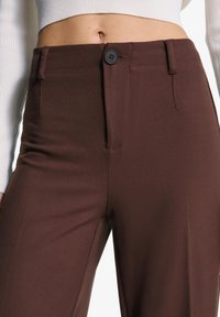 Brown high-waisted trousers with a smooth texture, featuring a front button closure, functional pockets, and tailored fit detail.