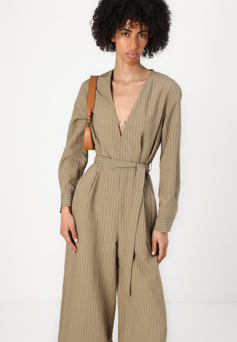 Beige gestreifter Jumpsuit mit tiefem V-Ausschnitt, langen Ärmeln und tailliertem Bund. Verfügt über eine glatte Textur und einen metallischen Akzent am Ausschnitt.