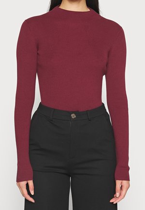 Femme portant un haut bordeaux moulant à manches longues en côtes, rentré dans un pantalon noir taille haute avec une fermeture boutonnée.