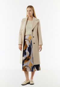 Manteau en laine beige avec revers et boutons noirs, superposé à une jupe à motifs bleus, orange et crème, associé à des chaussures slip-on beiges.