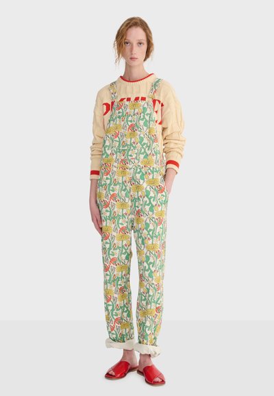 Jeune femme portant une salopette à motif floral, un pull crème avec des accents rouges, des manches retroussées et des sandales rouges à bouts ouverts, debout sur un fond blanc.