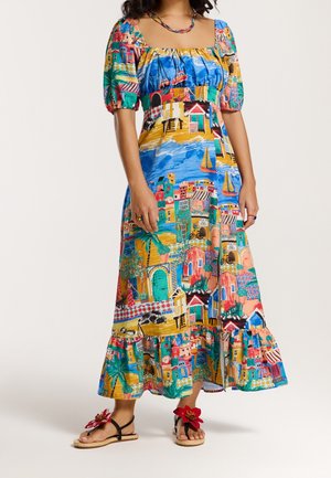 Mujer con un vestido midi colorido con mangas abullonadas y un estampado de pueblo costero, combinado con sandalias que tienen grandes decoraciones de flores rojas.