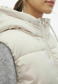 Gilet matelassé beige clair, avec un col montant et une fermeture éclair, offrant une texture douce et matelassée. La couche intérieure est grise et duveteuse.