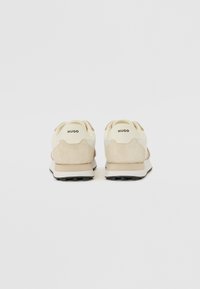Beige suede sneakers with white accents, featuring a raised back, textured fabric, and black rubber outsole, labeled “HUGO” on the heel.