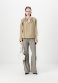 Blouse beige à manches longues avec un col en V, associée à un pantalon gris évasé. Le mannequin porte un petit sac à main blanc et des escarpins noirs à talons pointus.