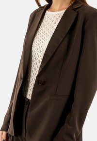 Blazer marrón hecho de tela suave, con un cierre de un solo botón y dos bolsillos frontales, combinado con una camisa blanca texturizada por debajo.