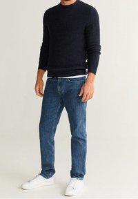 Uomo che indossa un maglione blu scuro, jeans blu e sneaker bianche in piedi contro uno sfondo chiaro e neutro.