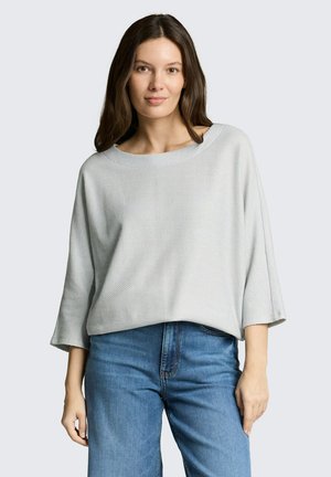 Vrouw met lang bruin haar, gekleed in een lichtgrijze gebreide trui en blauwe spijkerbroek, staand tegen een effen achtergrond.