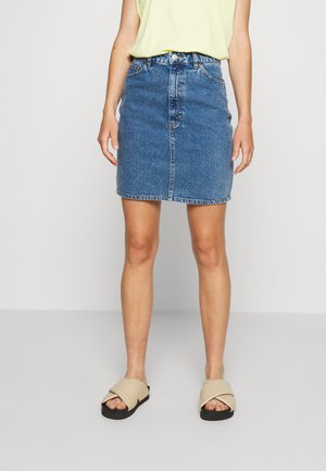 Woman wearing a blue denim knee-length skirt, light yellow sleeveless top, and beige slide sandals with black soles.