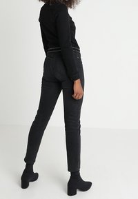 Persona vistiendo jeans ajustados negros, cinturón negro con tachuelas, chaqueta negra y botines negros de tacón, de pie contra un fondo blanco.