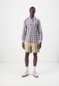 Plaid button-up shirt in red, white, and blue, khaki shorts, white socks, and tan sneakers with green accents. Model stands on a white background.