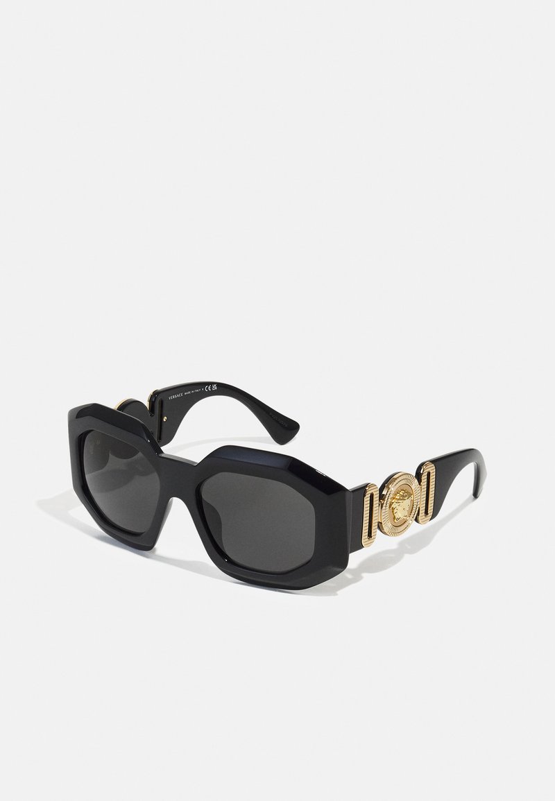 Occhiali da sole neri oversize con una montatura ottagonale, lenti scure e dettagli in oro con un emblema decorativo sulla tempia.